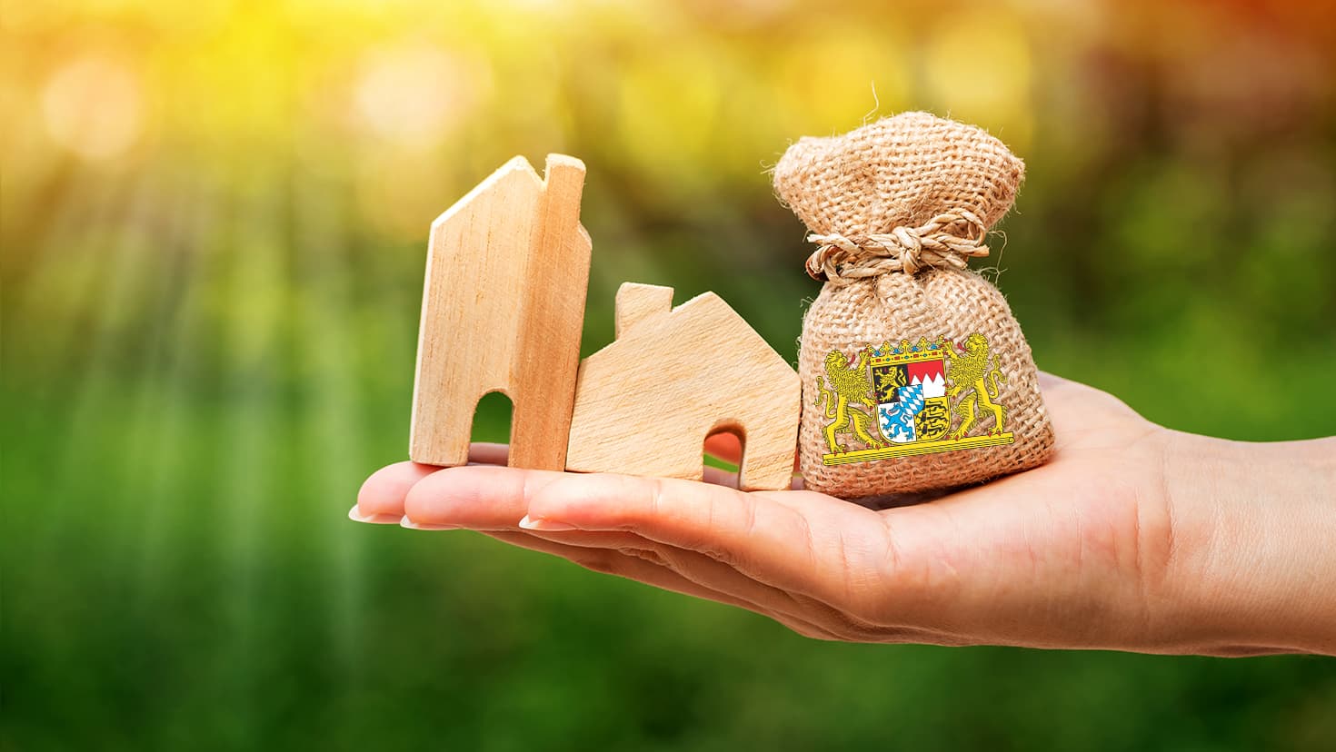 Hand hält Geldbeutel (links) und ein Musterhaus (rechts). Auf dem Geldbeutel ist das bayerische Staatswappen. © Watchara - stock.adobe.com