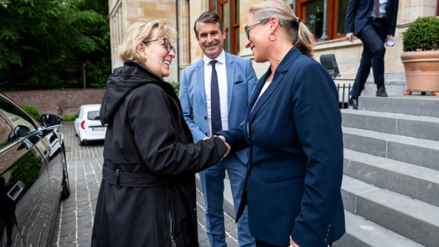 Die beiden Damen sch&uuml;tteln sich l&auml;chelnd die Hand. Minister Bei&szlig;wenger ist in der Mitte zu sehen und l&auml;chelt. Rechts geht es die Treppe hinauf.